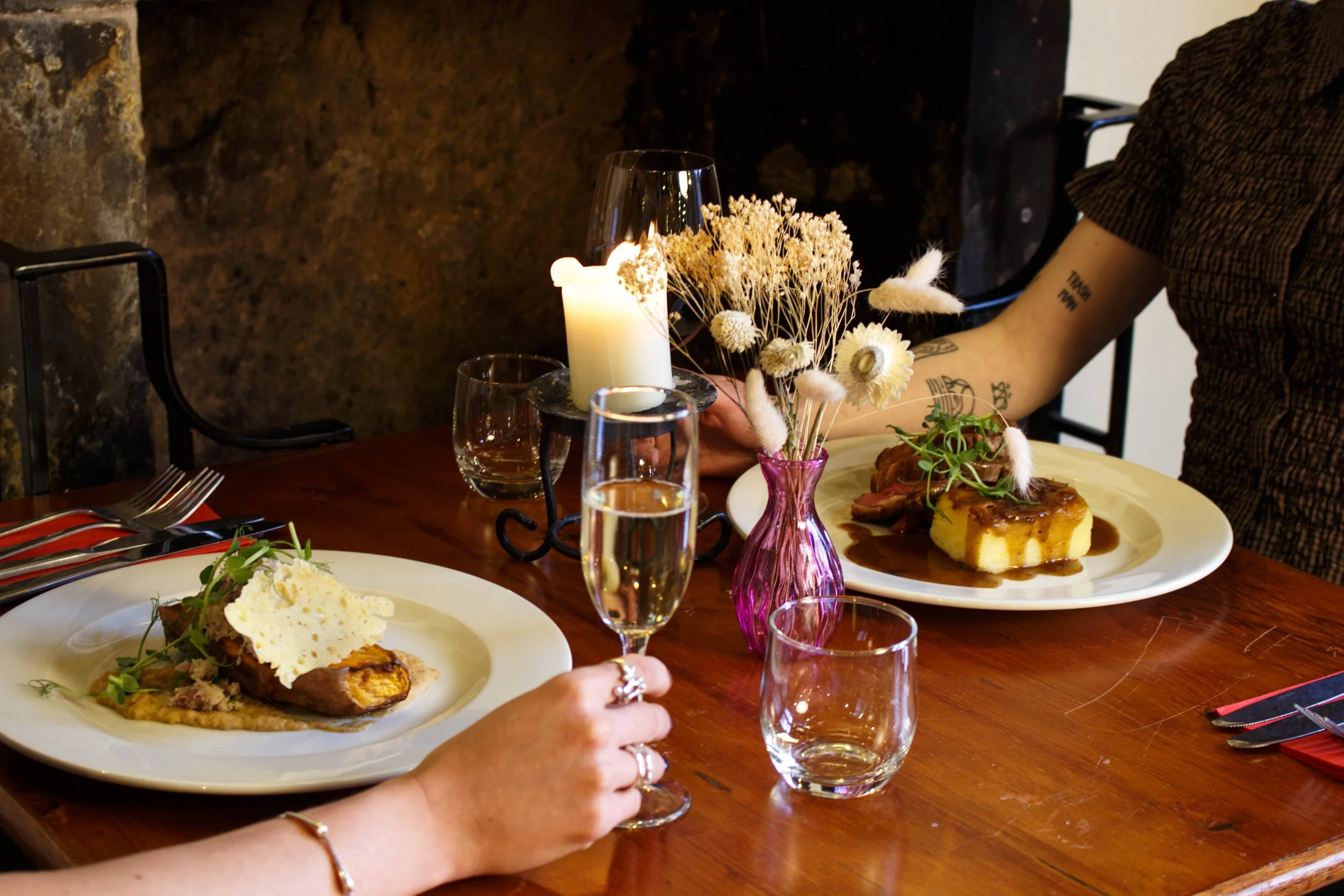 A dining table set with plates of food, wine glasses, and a centerpiece of dried white flowers in a pink vase. There is a candle, a glass of water, and a person with tattoos wearing a brown sweater reaching for food.