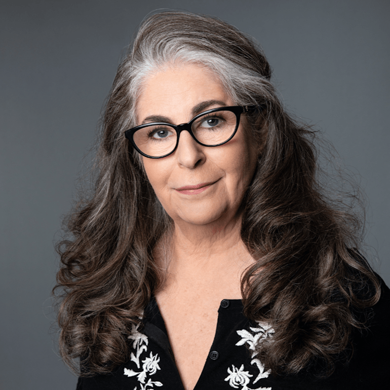 Sherry Salman, Jungian analyst and author of Oracle of Rewilding and Dreams of Totality. A middle-aged woman with long, wavy gray hair and black-framed glasses, wearing a black shirt with white embroidery, against a gray background.
