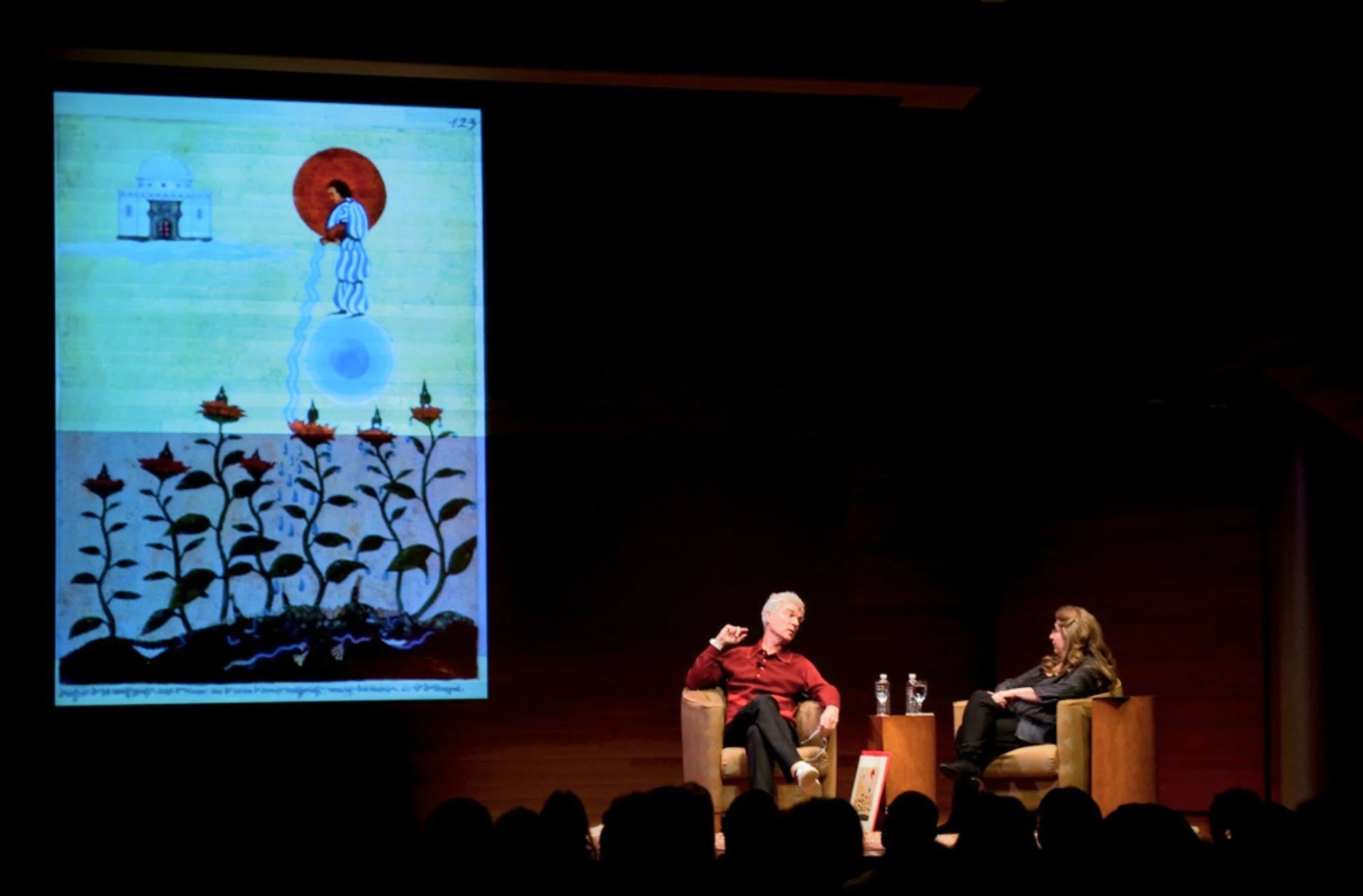 A panel discussion on stage with David Byrne and Sherry Salman in The Red Book Dialogues at the Rubin Museum of Art in New York City. A large painting is projected on the wall behind them.
