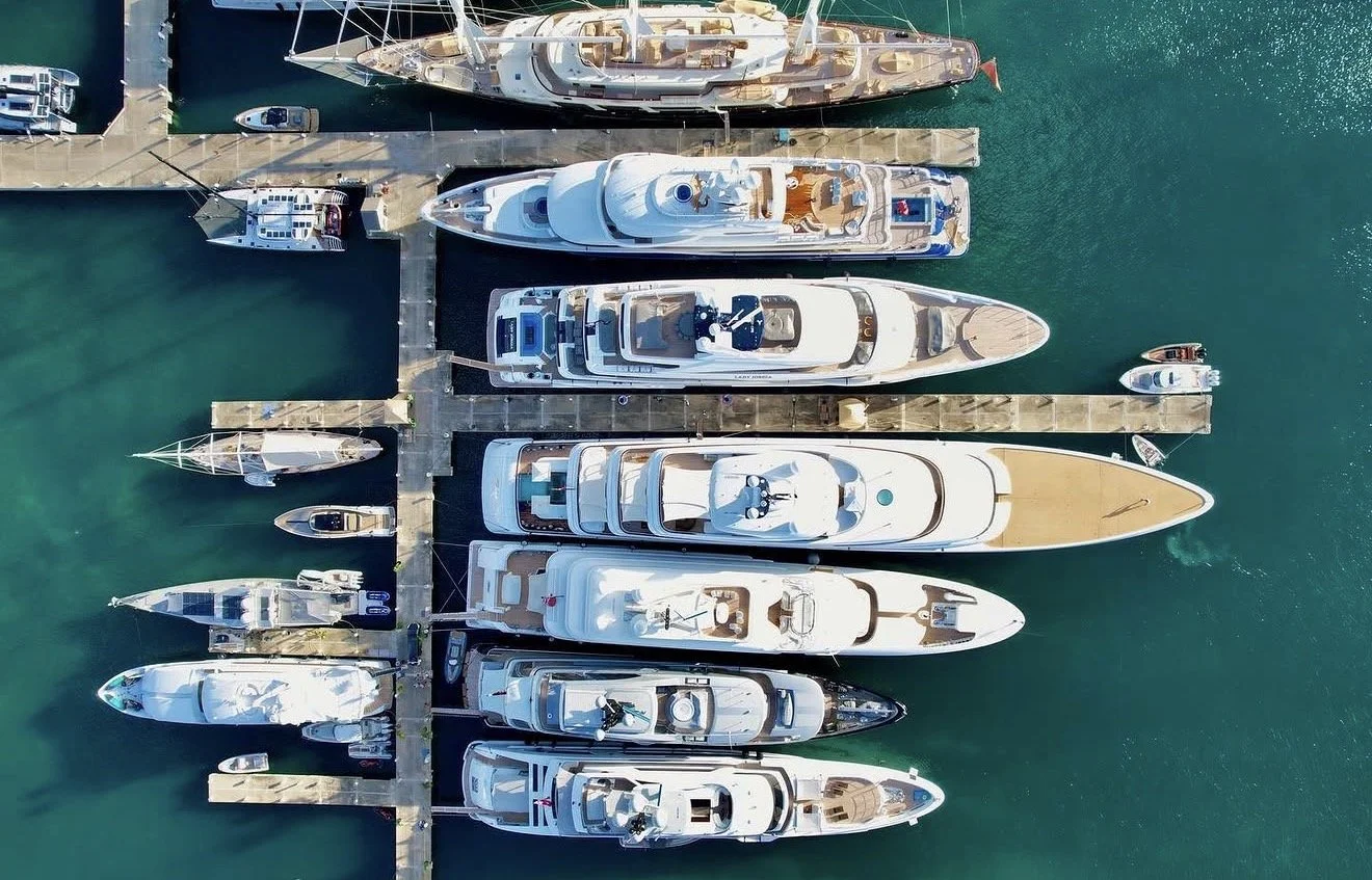 Overhead view of a marina with multiple large yachts docked alongside each other, with smaller boats and water visible around the pier.