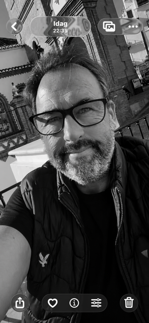 A man with glasses and a beard taking a selfie outdoors. There are buildings and a decorative gate in the background.