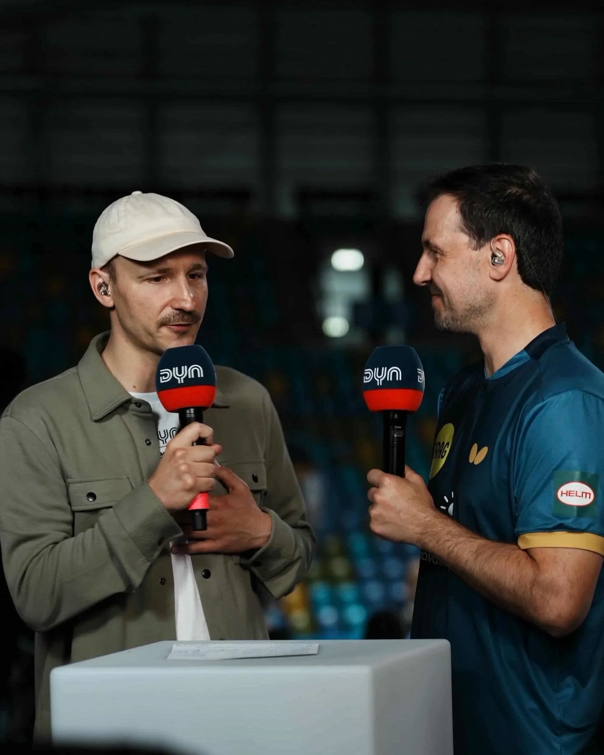 Zwei Männer mit Headsets interviewen ein Kind, das an einem weißen Podium steht, im Hintergrund ist eine dunkle, unscharfe Halle zu sehen.