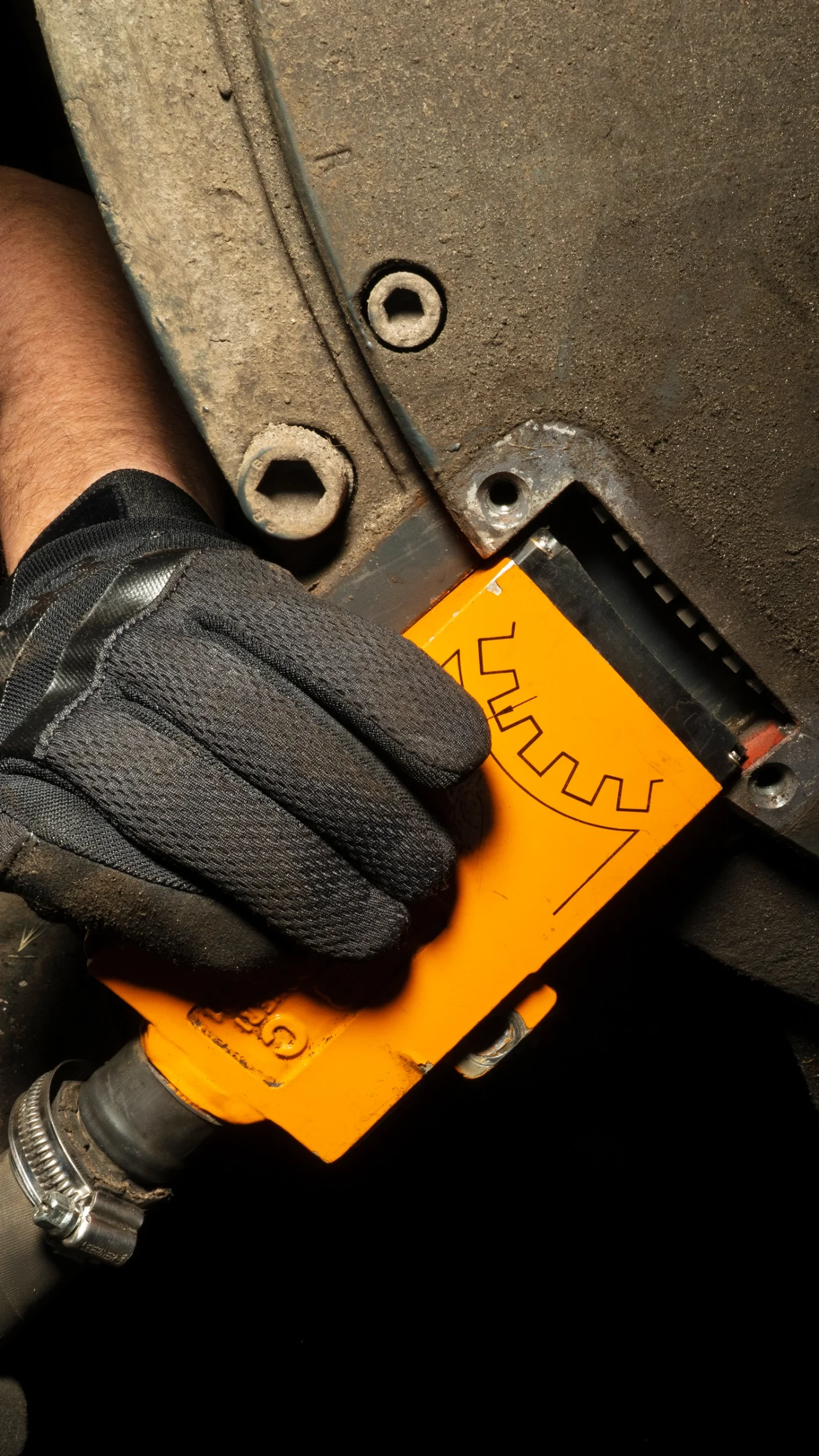 Eine Person trägt eine schwarze Arbeitshandschuh und hält ein orangefarbenes Messgerät mit einem schwarzen Kabel, das an eine Metallmaschinenteile angeschlossen ist.