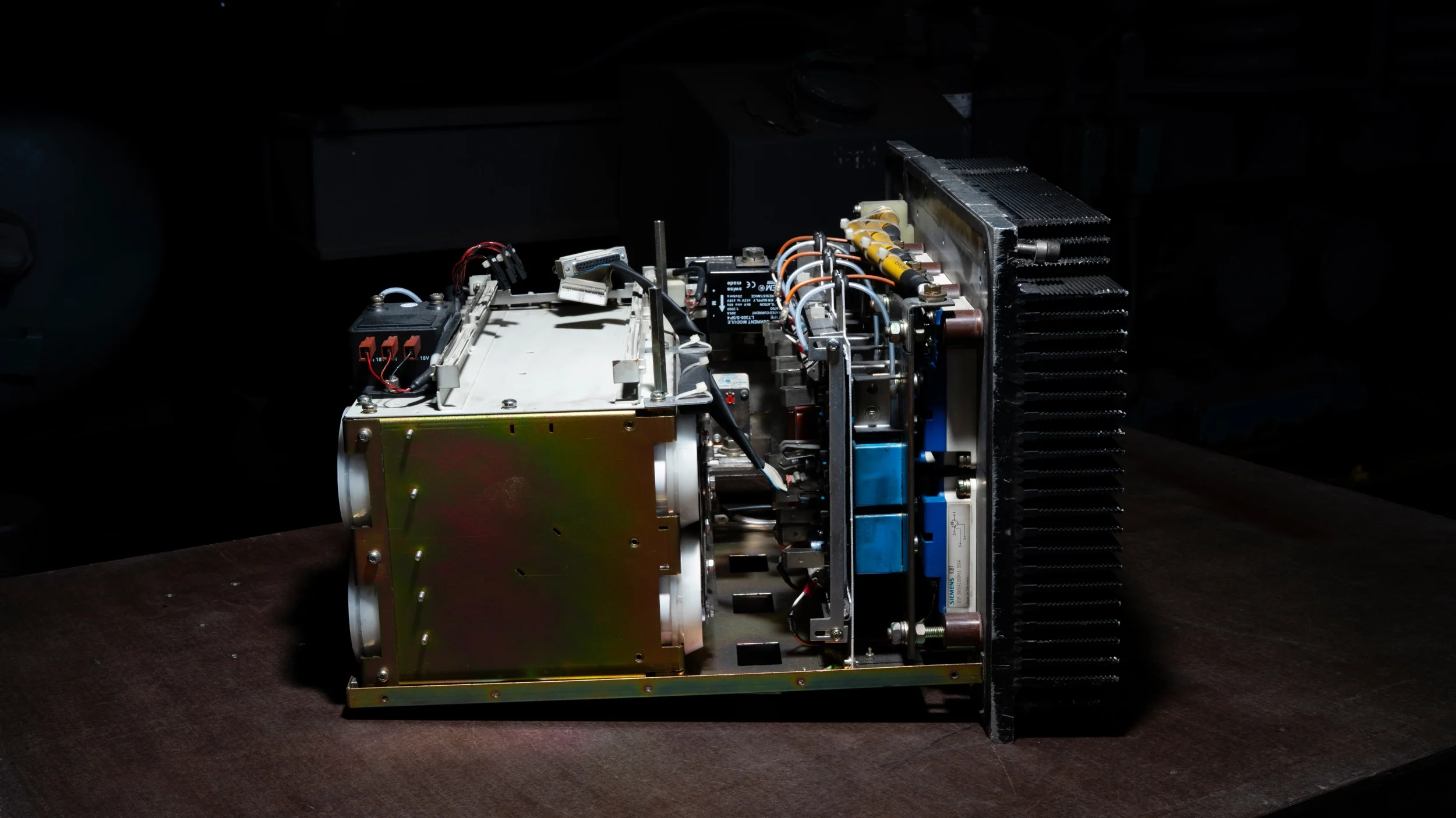 Nahaufnahme eines technischen Geräts mit mehreren Komponenten, Kabeln und Metallteilen, auf einem dunklen Hintergrund, vermutlich eine Elektronikeinheit oder ein Computerteil.