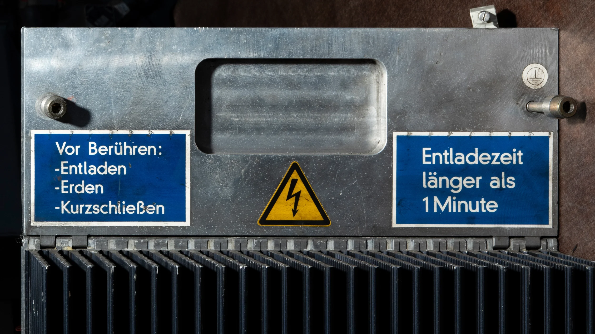 Metallgehäuse mit Warnsymbol, blaue Hinweise auf Entladezeit und Sicherheitsmaßnahmen, einschließlich Erdung und Schutzvorrichtungen.