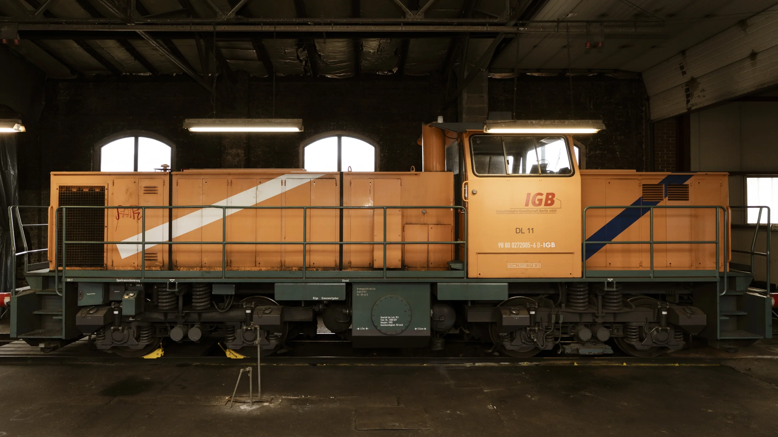 Ein gelber Güterwagen mit blauen und weißen Streifen, marked mit 'IGB', steht in einem Industriegebäude mit schwarzen Wänden und drei großen Fenstern im Hintergrund.