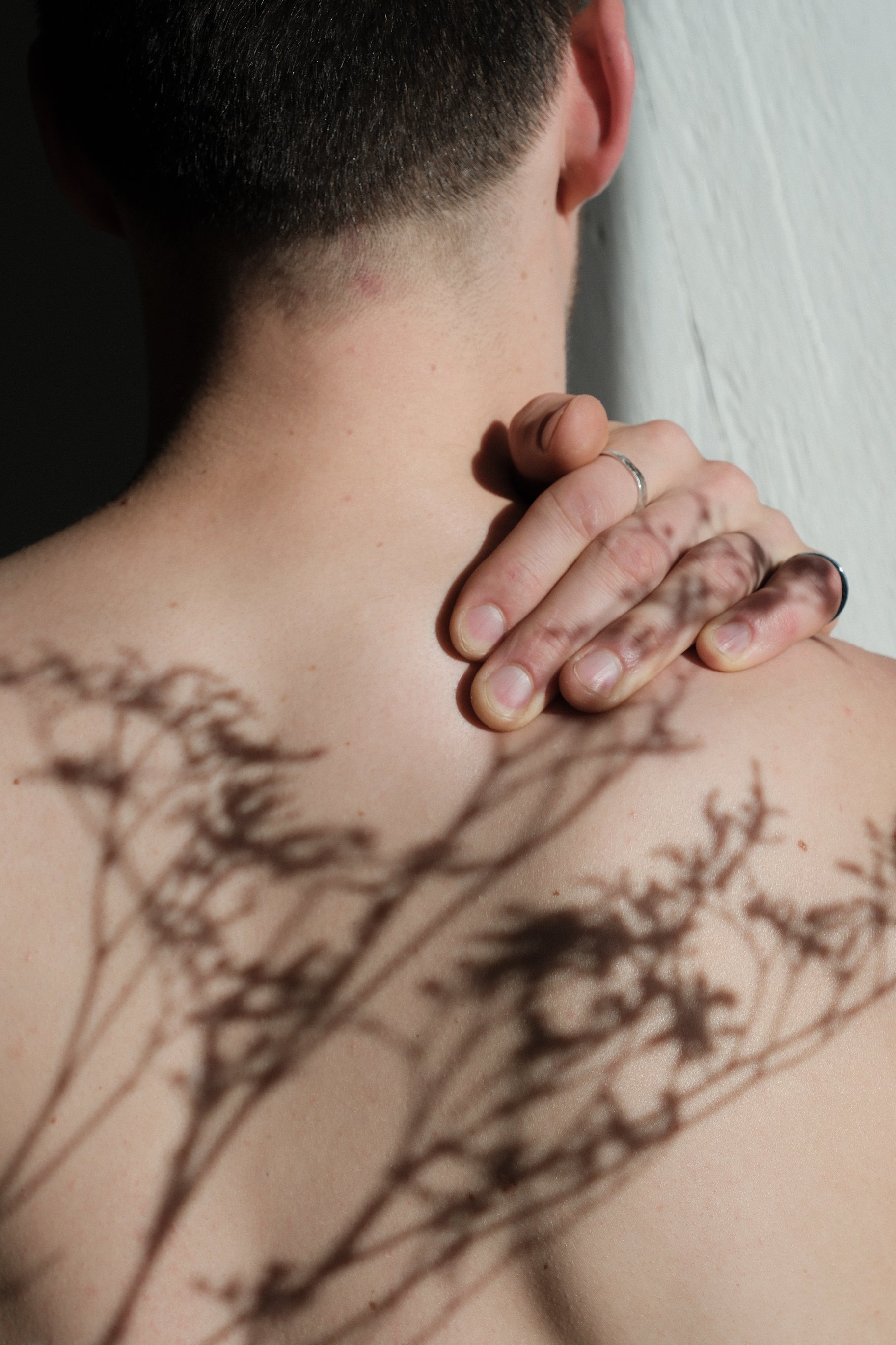 Nahaufnahme eines Menschen, der den Hinterkopf und das Nackenbereich zeigt, mit einer Hand, die den Hals berührt und einem Schattenspiel, dass durch die Sonne und eine Blume entsteht.