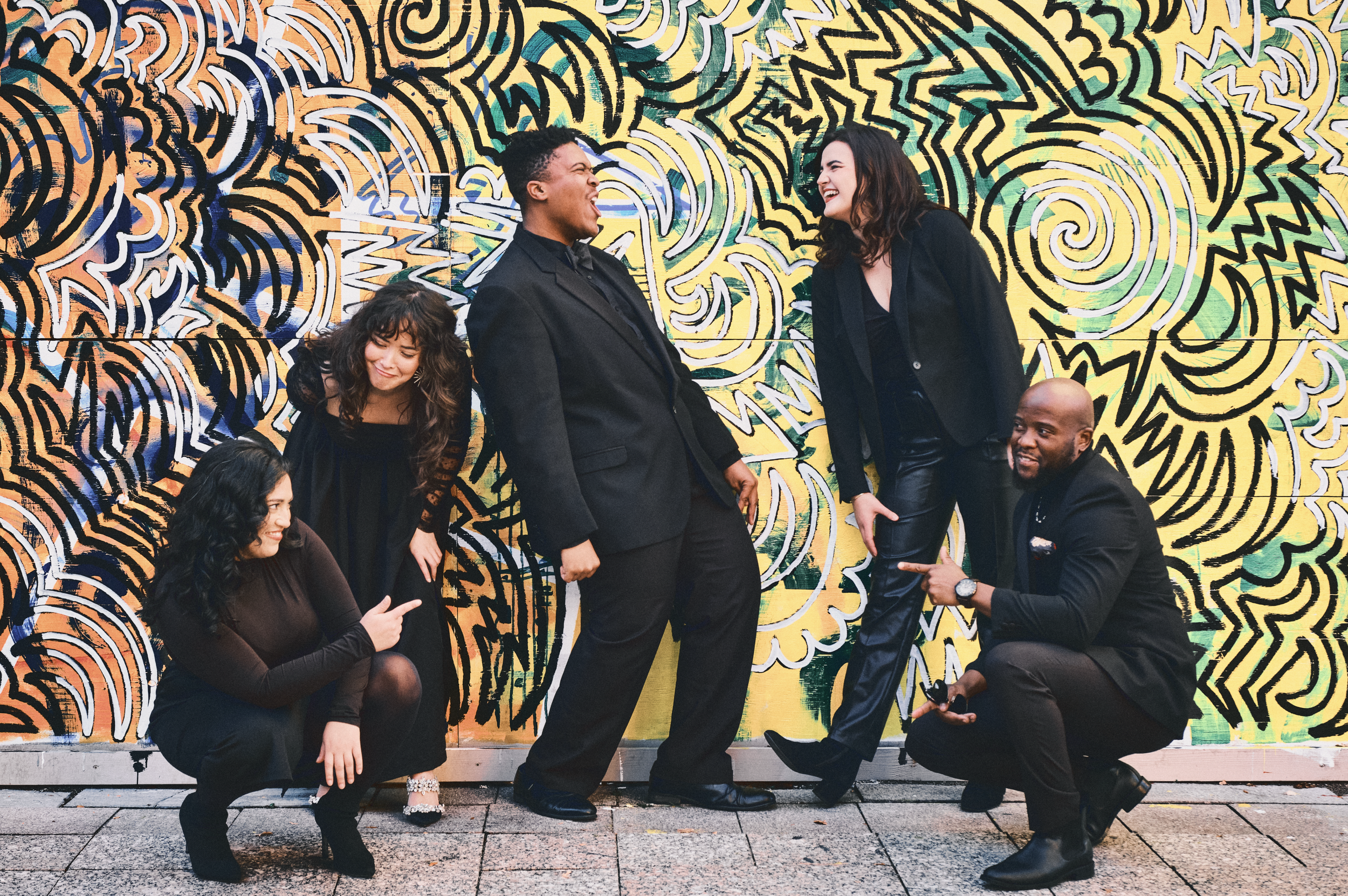 Five people interacting in front of a colorful graffiti wall. They are smiling, laughing, and pointing at each other, dressed in black formal and casual attire.