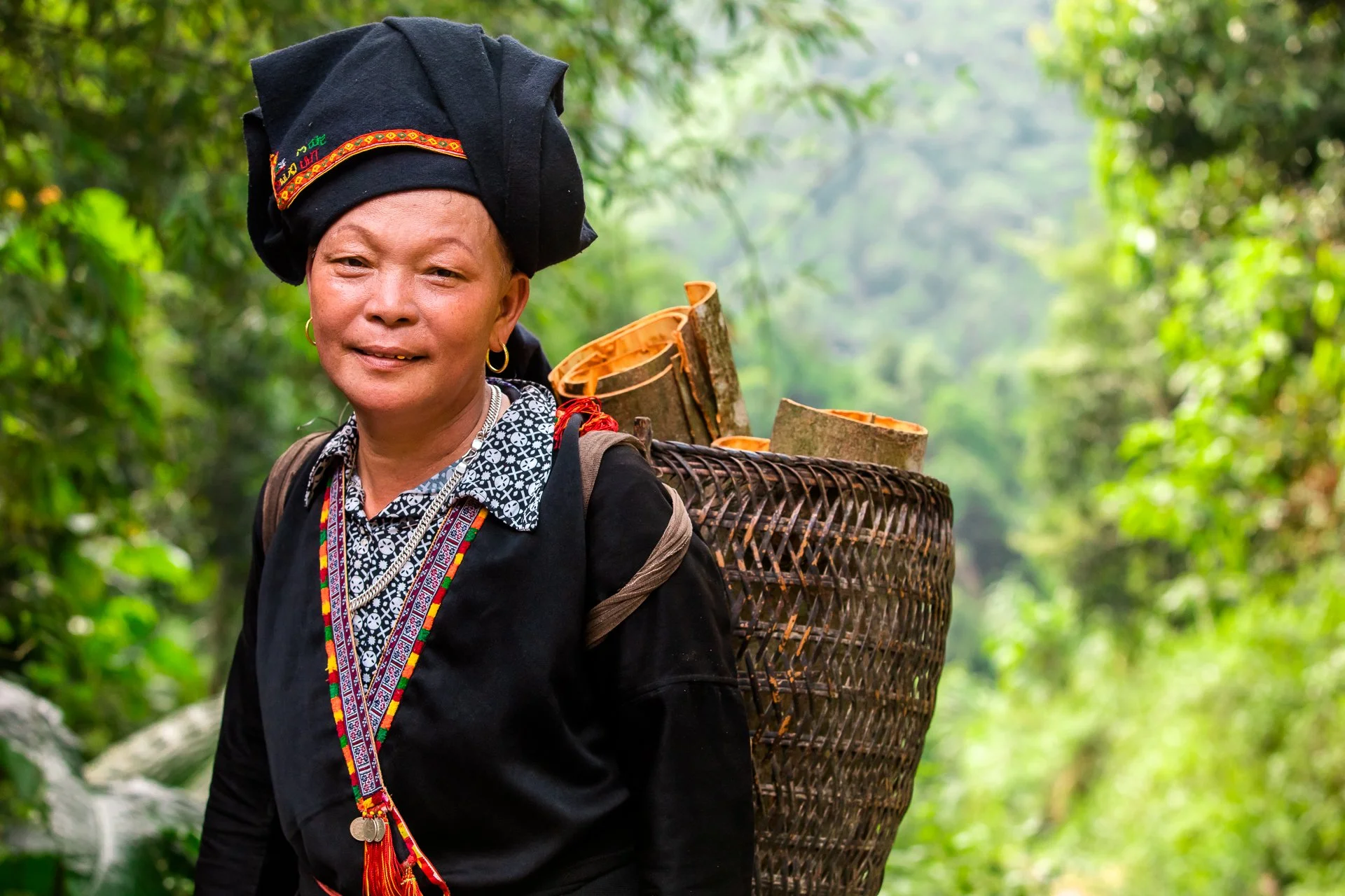 An indigenous woman in traditional attire carrying a basket on her back in a lush green forest.