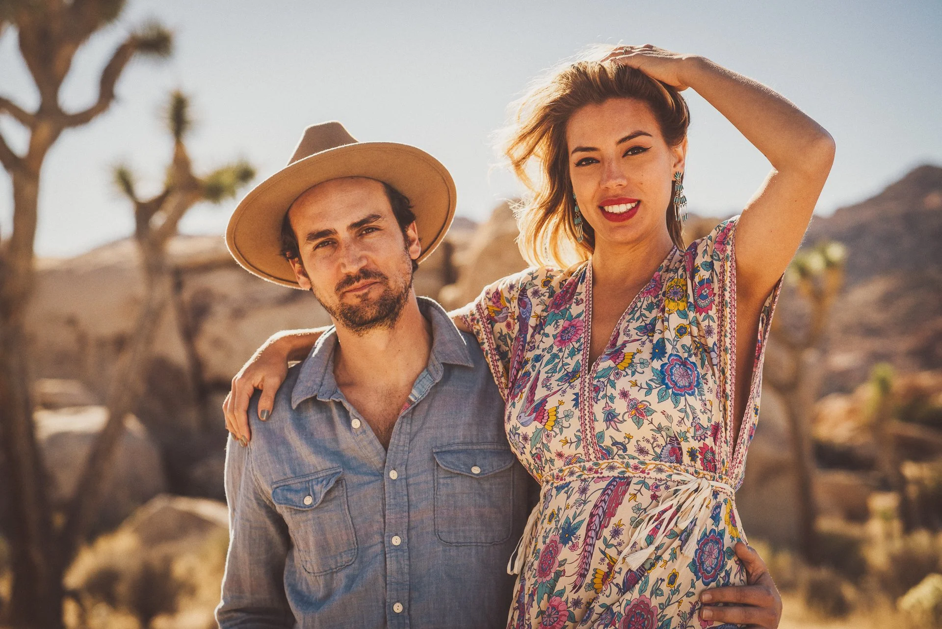 A man and a woman standing outdoors with desert and Joshua trees in the background, smiling at the camera.