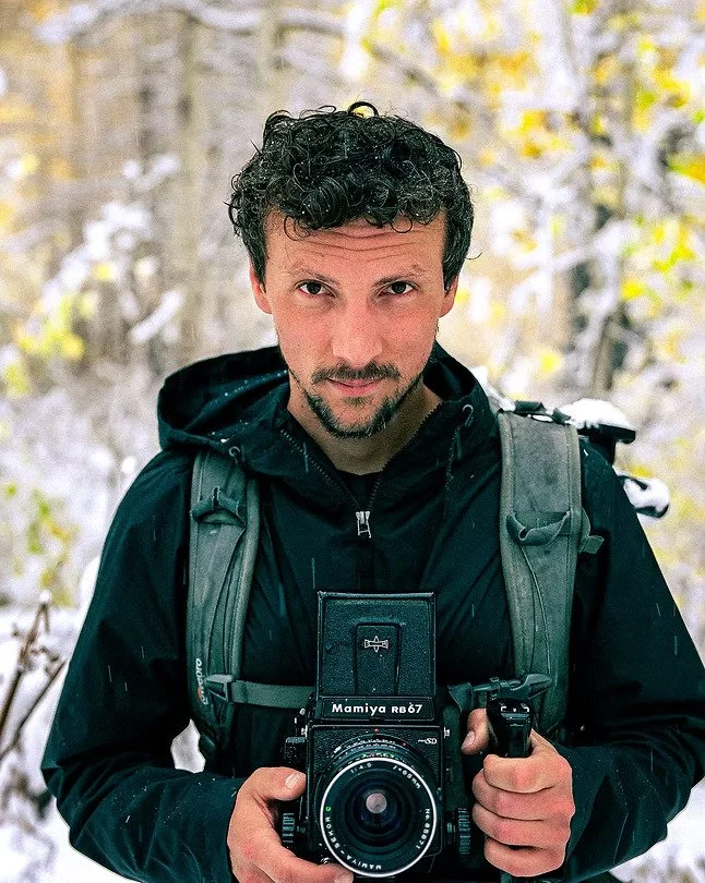 A man with curly dark hair and facial hair taking a selfie outdoors in a snowy forest, wearing a black jacket and carrying a camera and backpack.