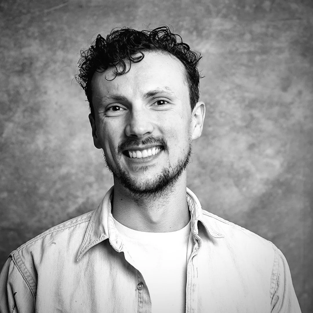 Black and white portrait of a smiling young man with curly hair, wearing a light-colored shirt and a white t-shirt underneath, against a textured background.