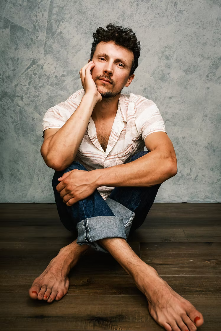 A man with curly dark hair, dressed in a light striped shirt and jeans, sits on a dark wooden floor against a textured gray wall, resting his chin on his hand and looking at the camera.