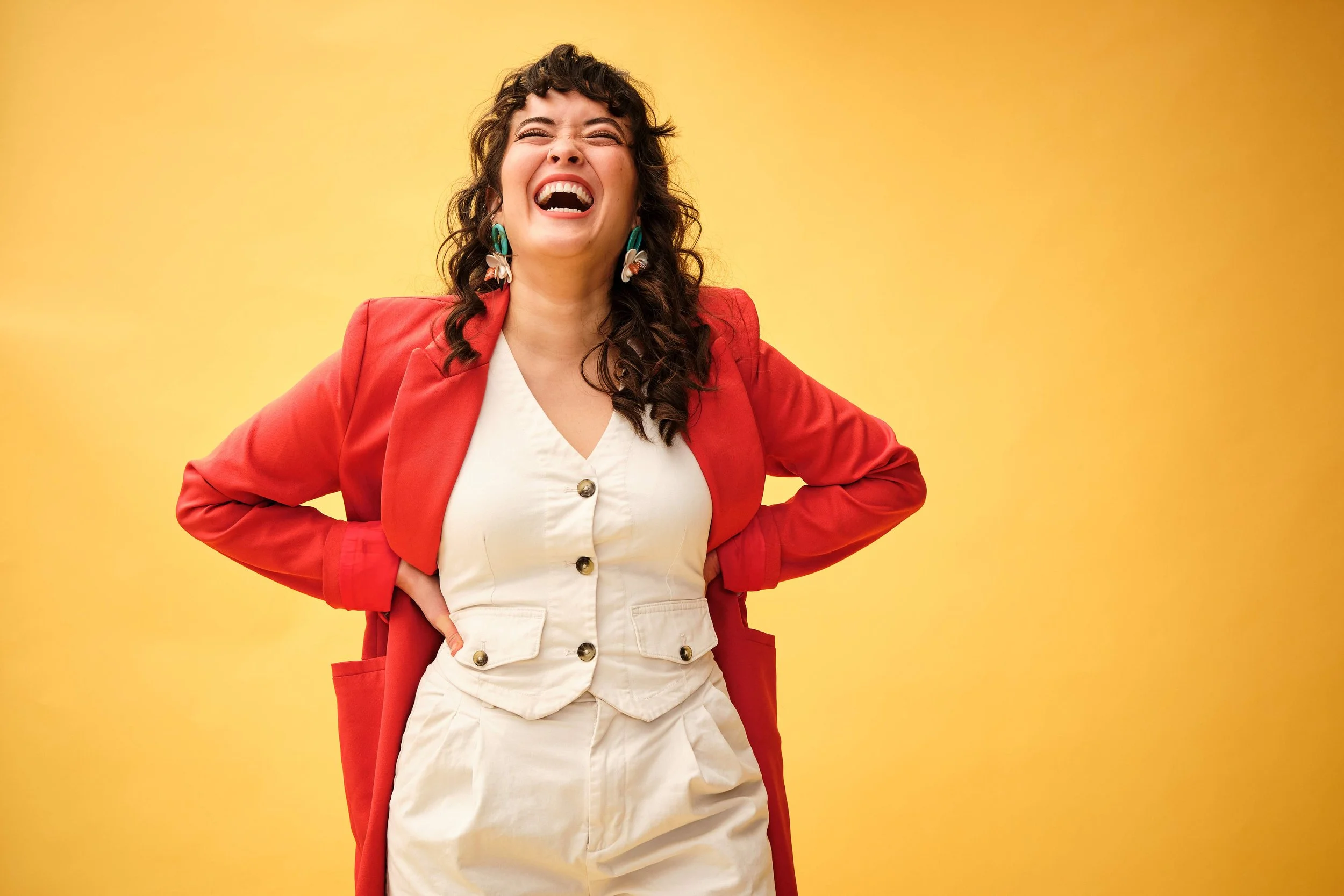 A woman with curly brown hair wearing a red blazer and cream-colored overalls, laughing against a yellow background.