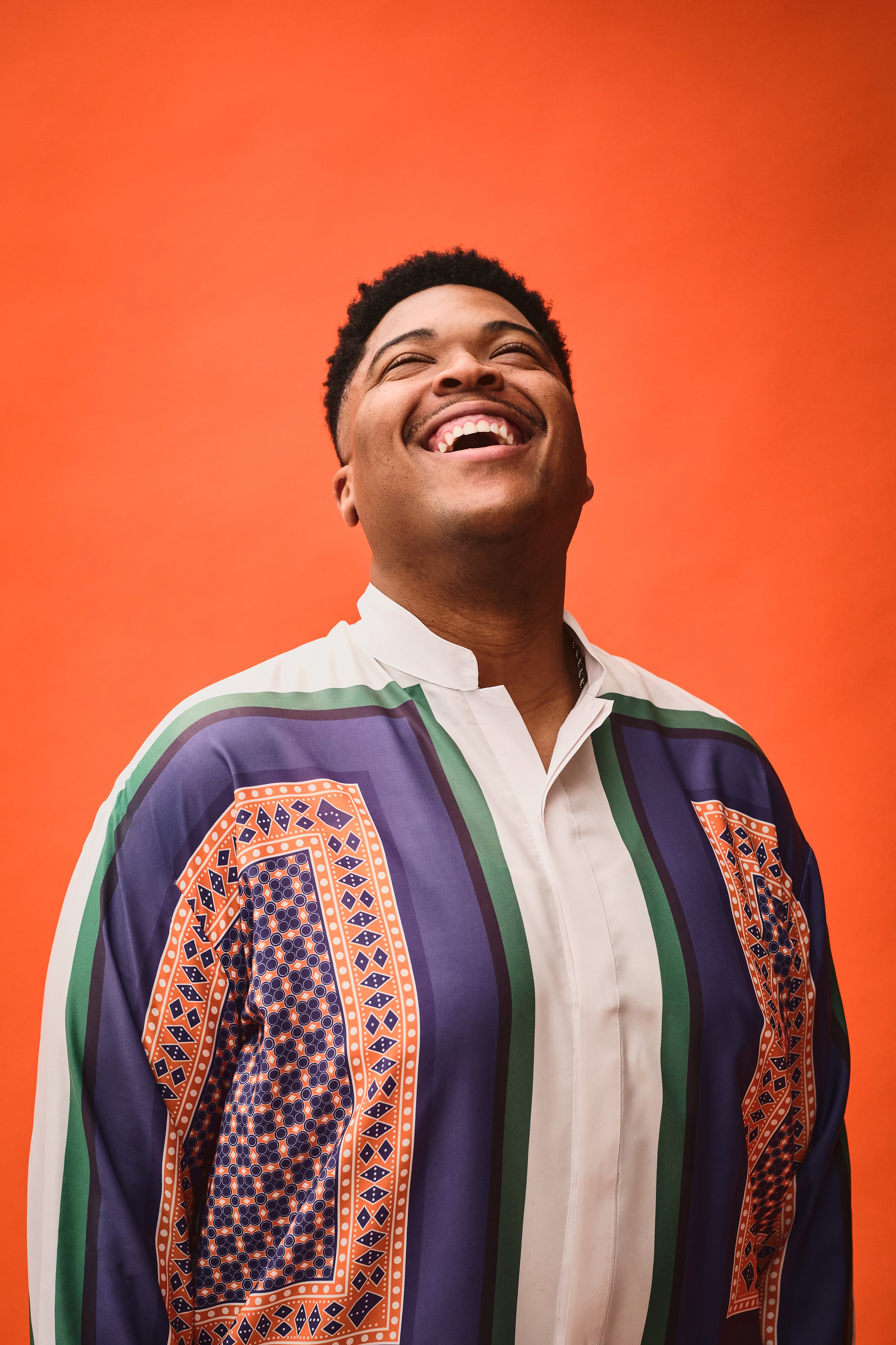 Person with short curly hair smiling, wearing a colorful patterned shirt, against an orange background.