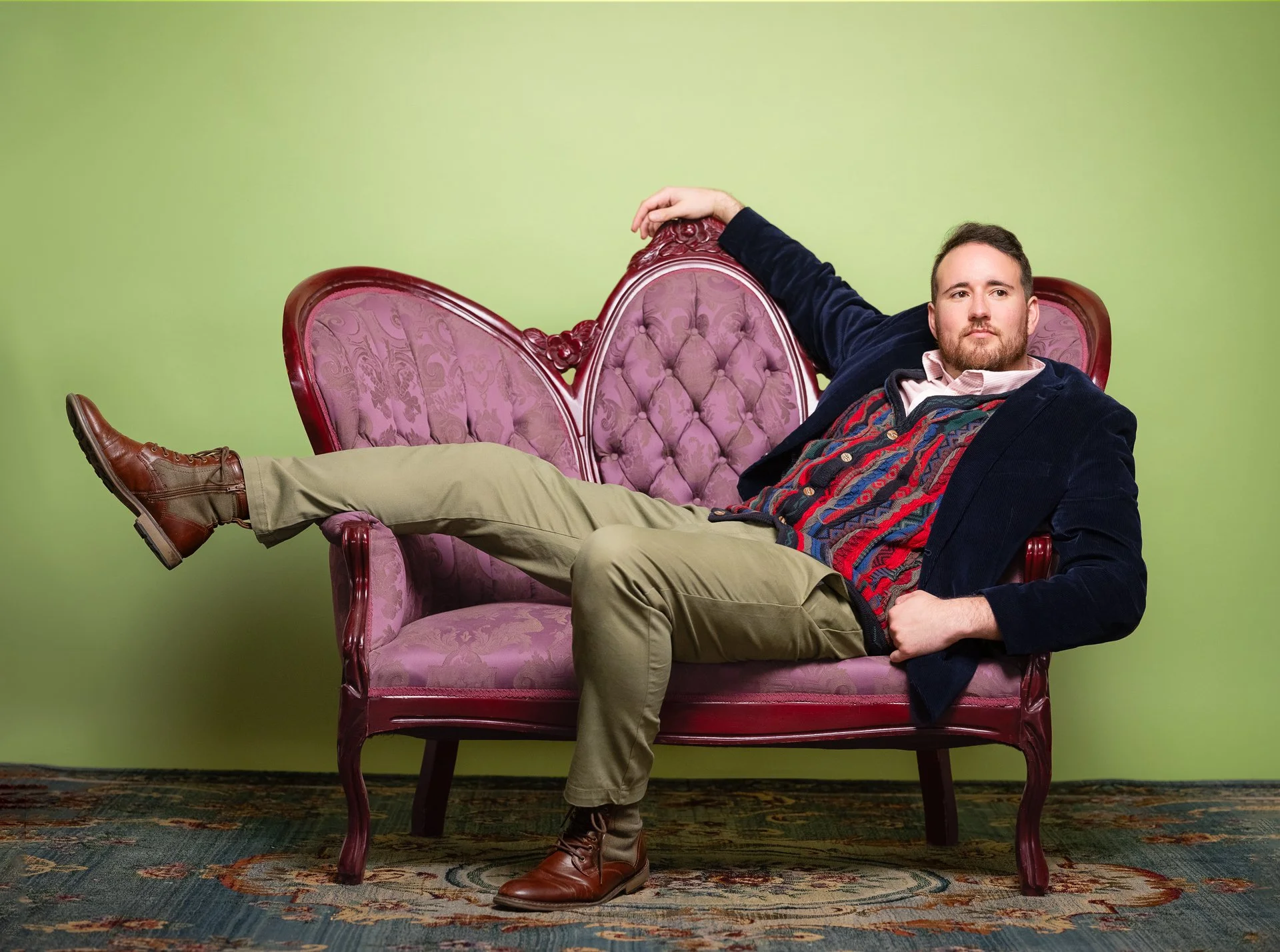 A man with a beard and short dark hair relaxing on a vintage pink upholstered settee with carved wooden frame, against a lime green wall. He's wearing a dark blazer, a colorful sweater, beige pants, and brown boots, with one arm draped over the back 