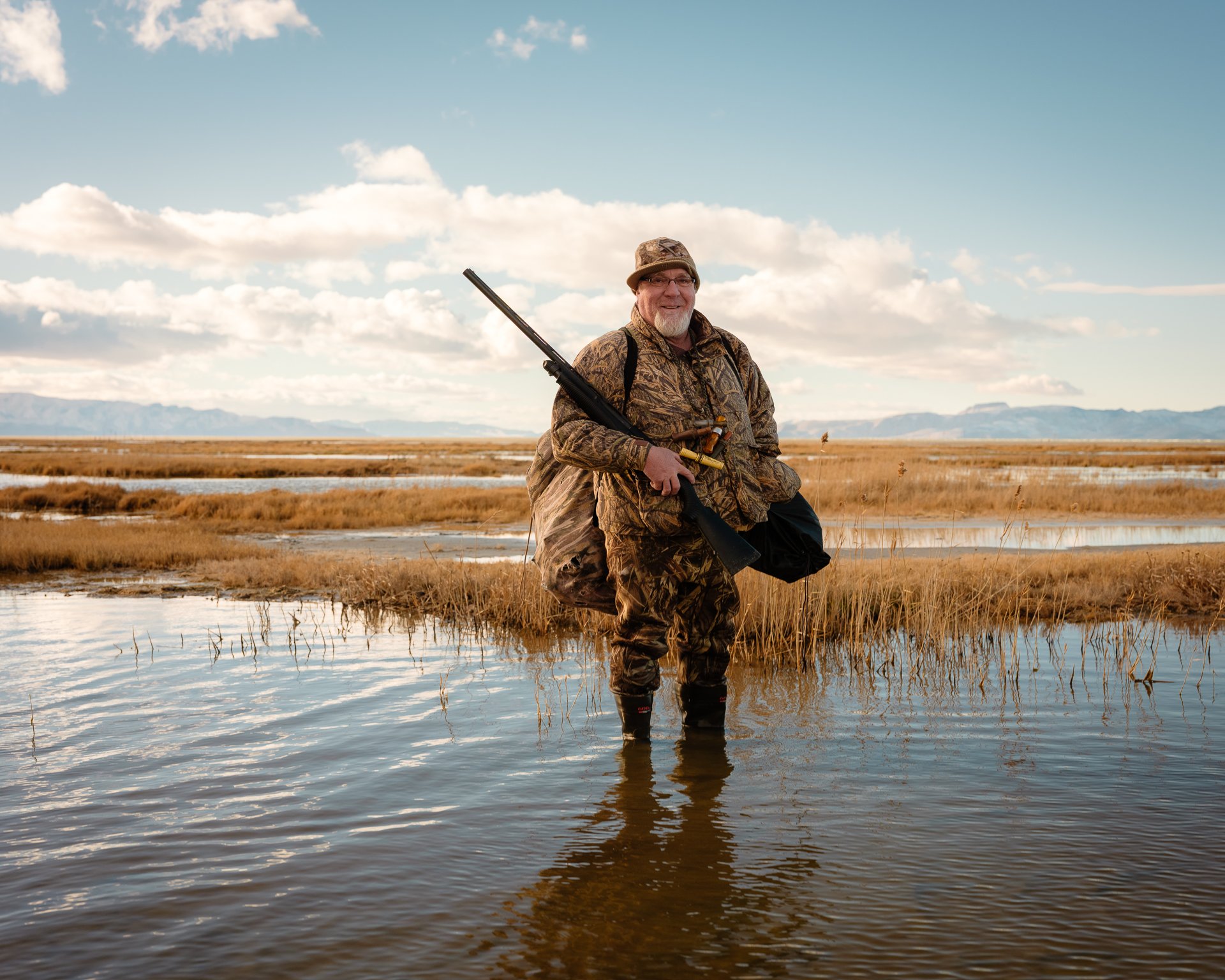 A man in camouflage clothing stands in shallow water with a rifle slung over his shoulder, in an open marshland landscape with mountains in the distance under a blue sky with scattered clouds.