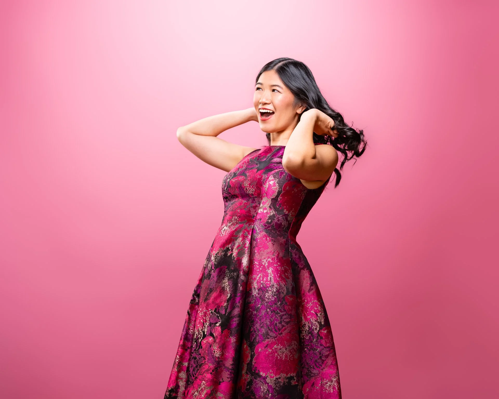 A woman with long black hair wearing a pink and black floral dress, smiling and posing with hands behind her head against a pink background.