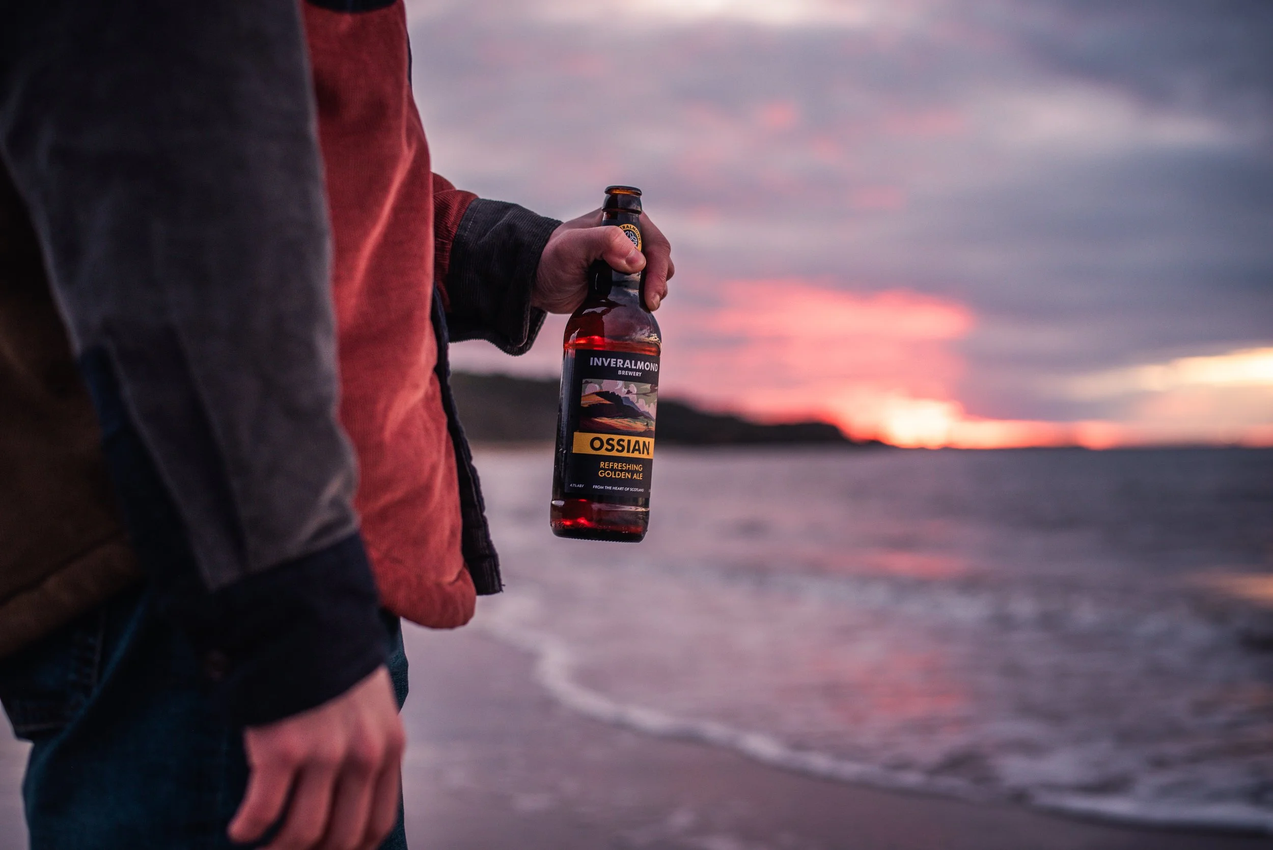 Person wearing a red jacket holding a bottled beverage labeled Ossian at sunset near water.