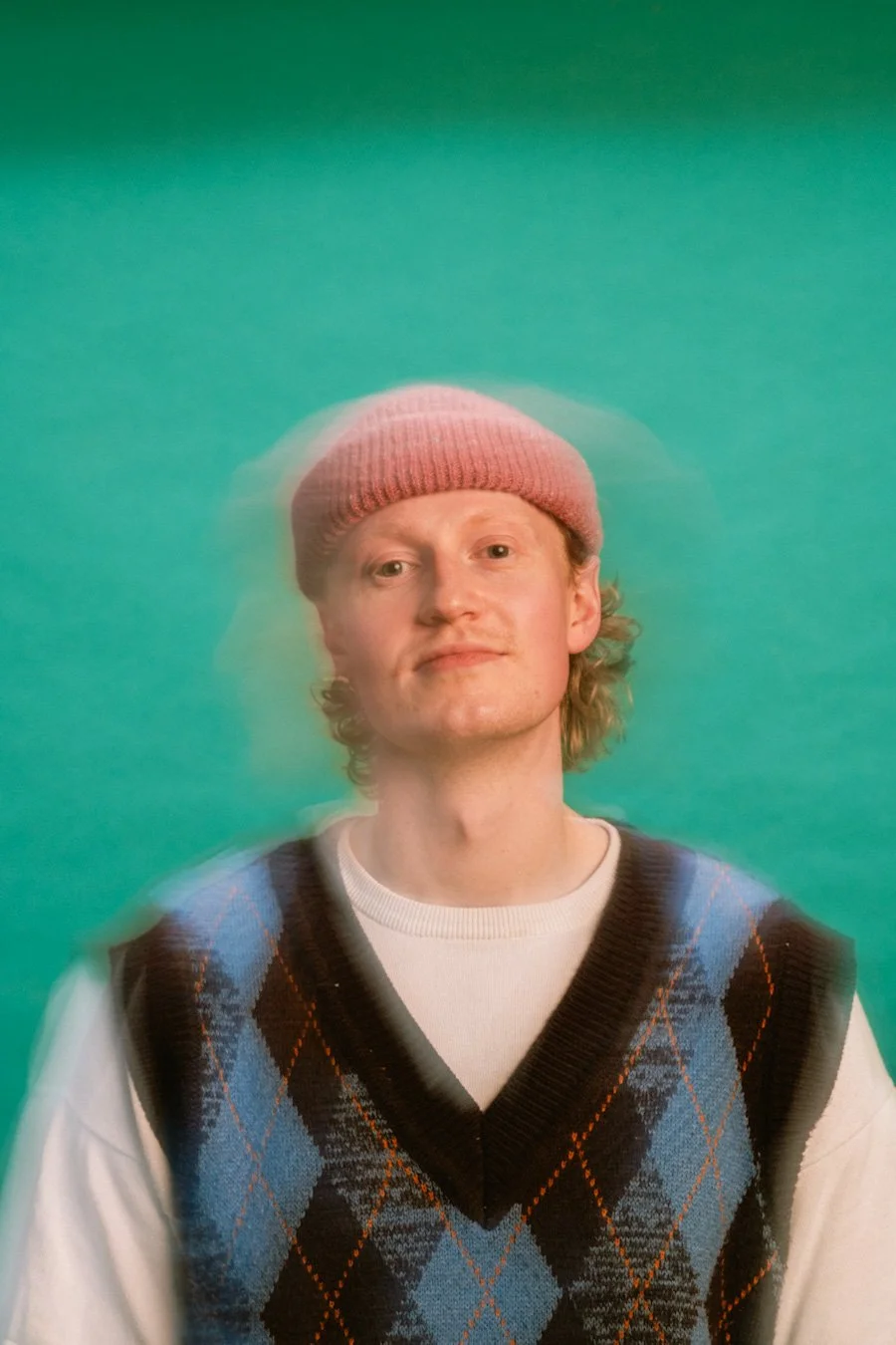 A young man with curly blond hair wearing a pink beanie, a white shirt, and a blue argyle sweater vest, standing against a green background.
