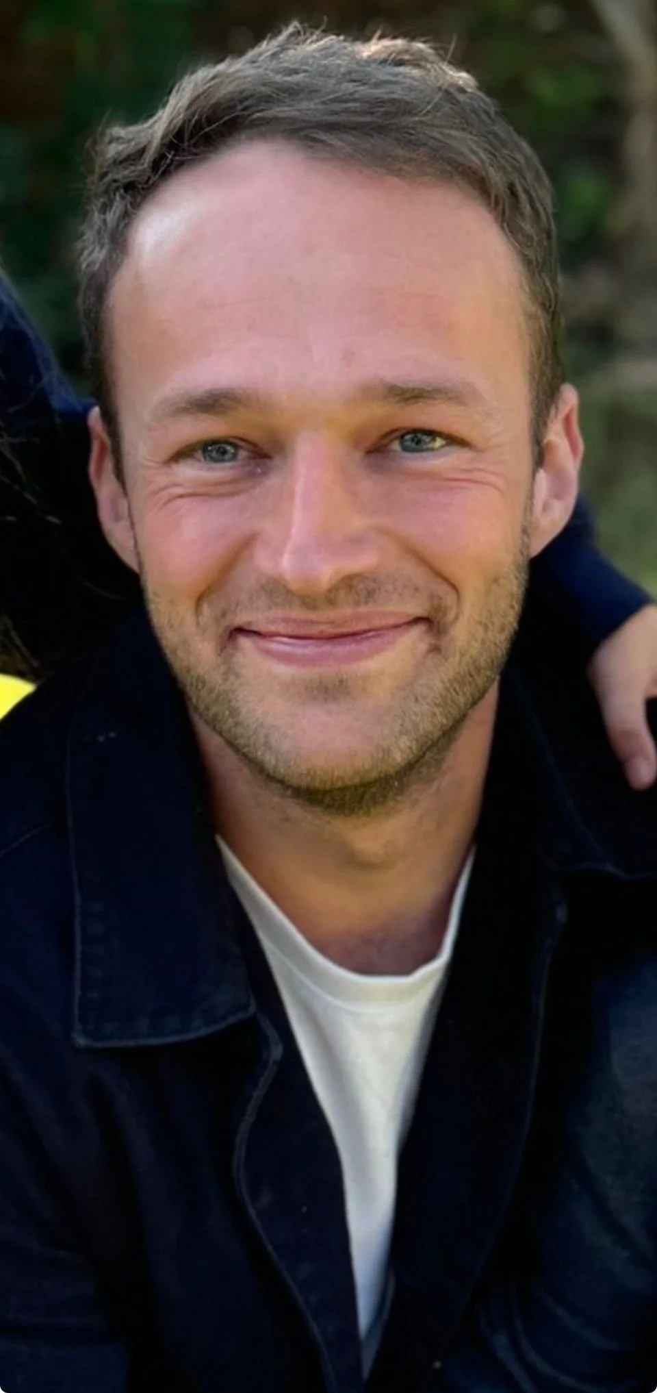 A smiling young man with short light brown hair and blue eyes wearing a black jacket and a white shirt, outdoors with greenery in the background.