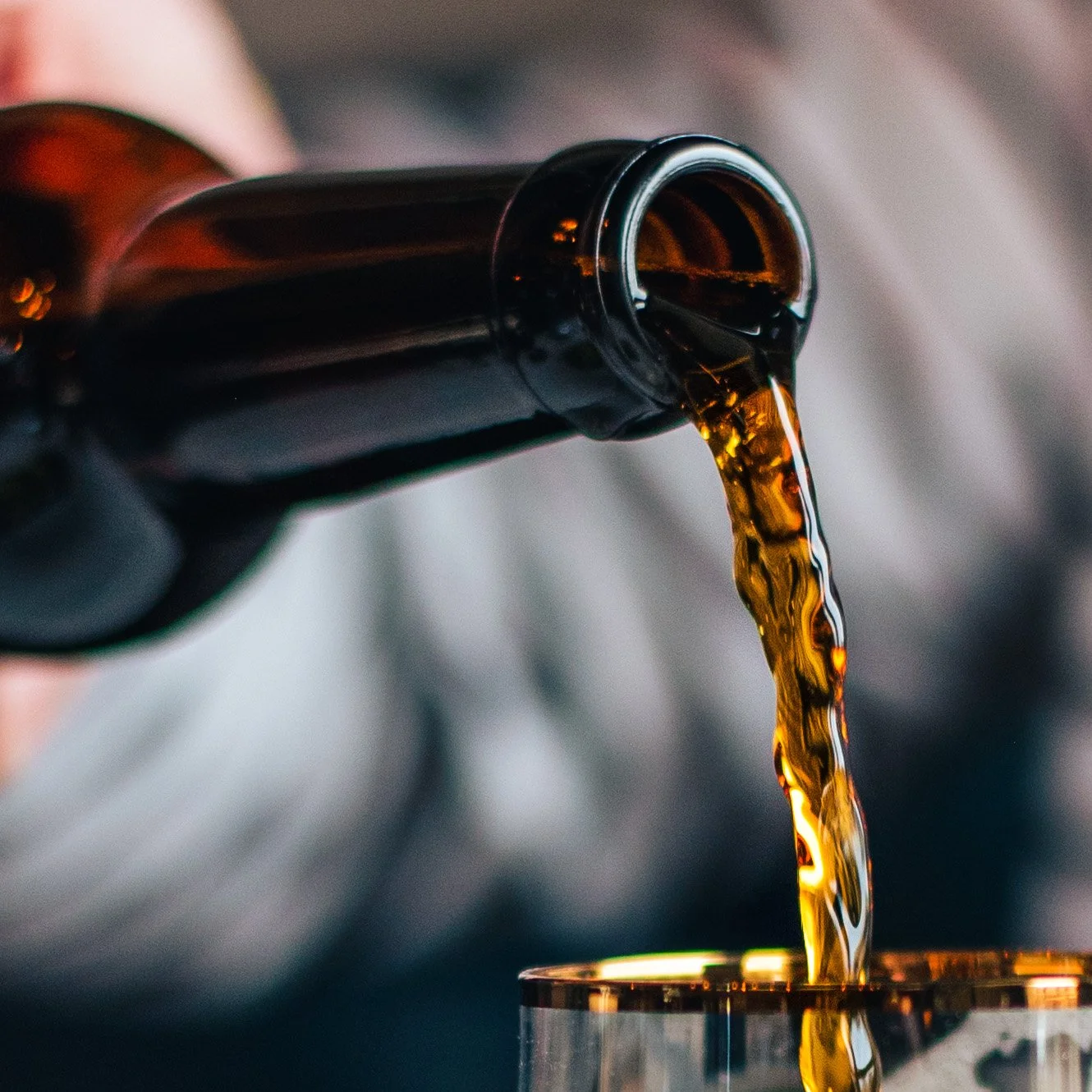 A hand holding a dark glass bottle tilted, pouring a dark liquid, possibly soda or beer, into a metal cup.