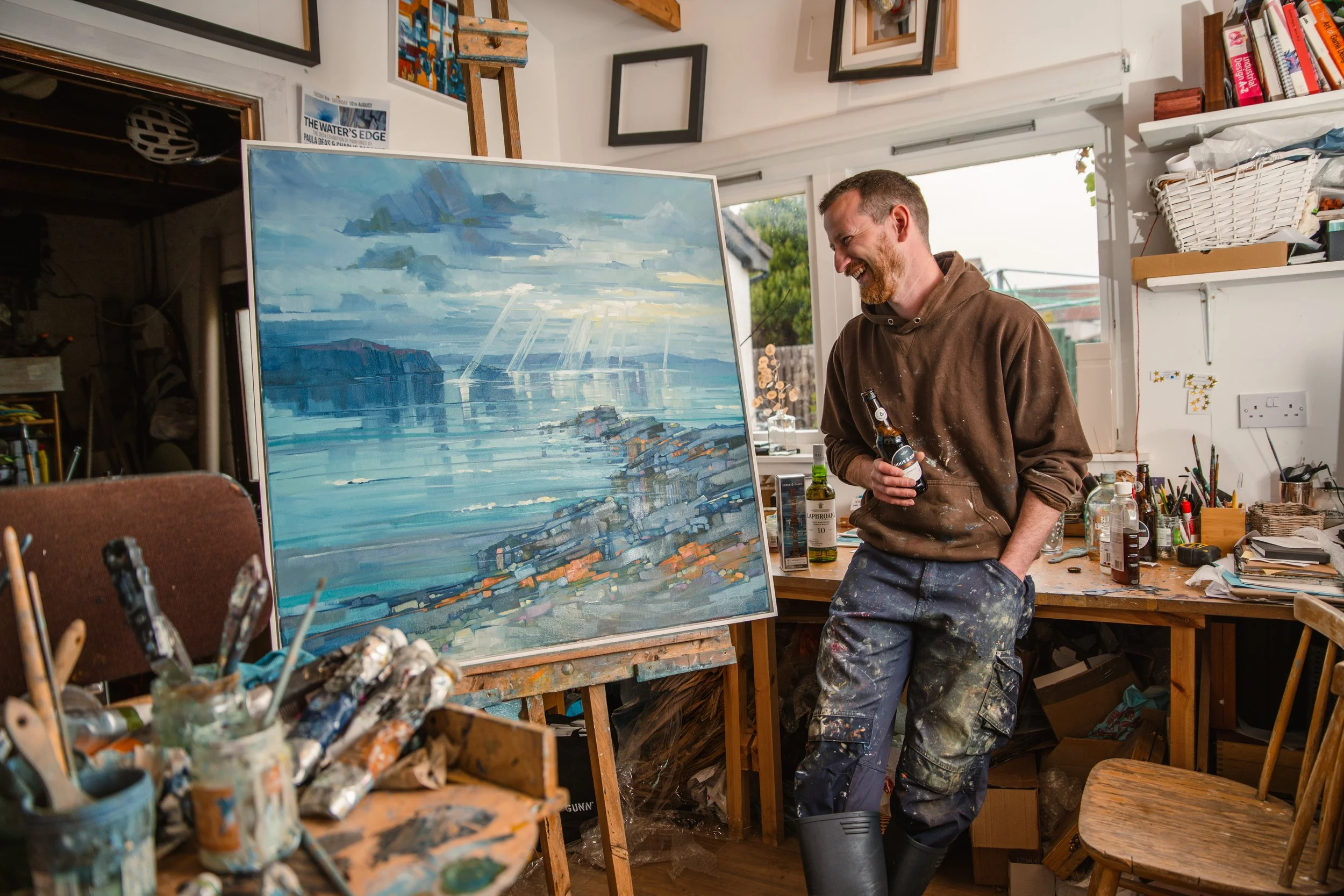 A man smiling in a brown hoodie and paint-stained pants stands in an art studio, holding a beer bottle while working on a large abstract landscape painting on an easel surrounded by art supplies and shelves of books.