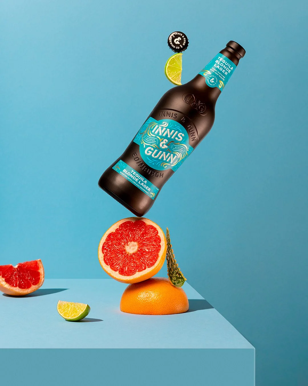 A bottle of Tequila & Gunny Blonde Lager beer appears to be floating above a halved grapefruit, a citrus wedge, and a clementine placed on a light blue surface against a blue background, creating a surreal, colorful arrangement.