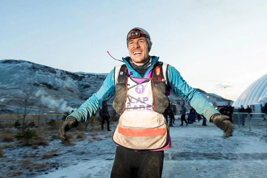 A male runner smiling in winter gear and headlamp, reaching the end of a race in a snowy landscape, with other people and a white dome structure in the background.