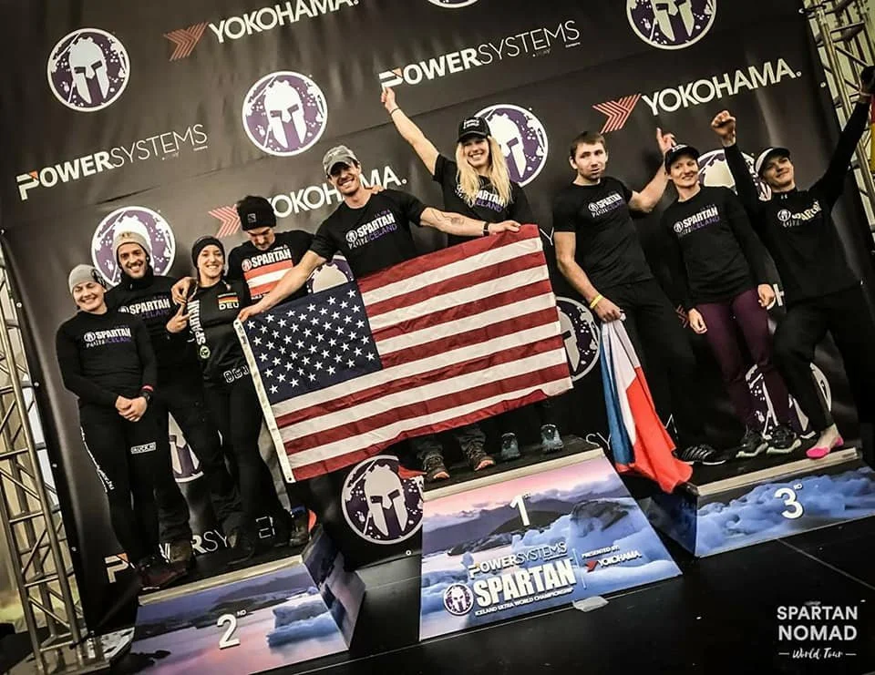 Group of eight people on a podium celebrating, holding an American flag, with a backdrop featuring the logos of Spartan and Yokohama. The first-place winners are in the center, smiling and raising their arms.