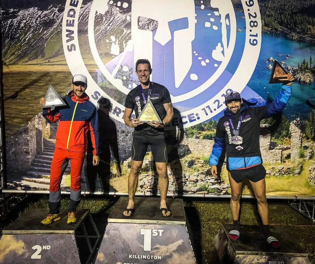 Three male trail runners on a winners' podium holding trophies, with a scenic outdoor backdrop featuring mountains and a lake. The man in the center is in first place, wearing shorts and flip-flops, holding a triangular trophy. The man on the left is in second place, dressed in a red and blue jacket, and the man on the right is in third place, wearing a black and blue jacket, holding a triangular trophy with a medal around his neck.