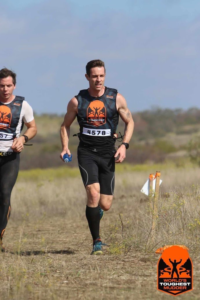 Two male runners participating in an outdoor race on a trail in a grassy field. The man in the front is wearing a black athletic outfit with a bib number 4578, holding a blue water bottle, and appears focused. The second man is partially visible and also dressed in athletic gear. There is a sign with an orange logo and the words "World's Toughest Mudder" on the bottom right corner of the image.