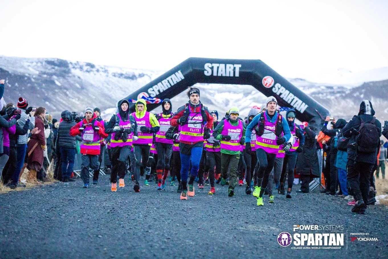 Group of runners at the starting line of a race under a black arch with 'START' and 'SPARTAN' written on it, in a mountainous, snowy landscape, with spectators and photographers along the sides.