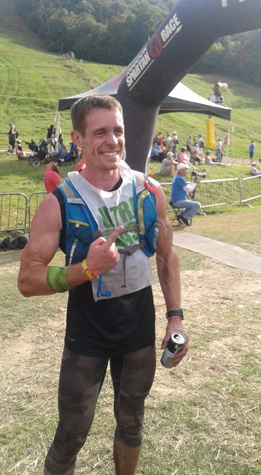 A smiling man in athletic gear holding a can of drink at a race event with a starting arch in the background.