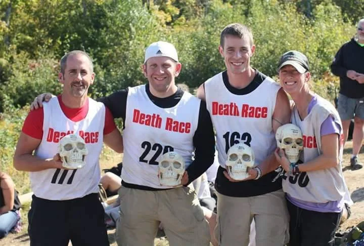 Four people holding skull trophies at a race called 'Death Race', smiling outdoors in a wooded area.