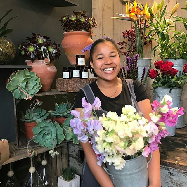 Young black woman smiling with a large bucket of colorful sweet pea flowers in a flower shop surrounded by plants and flowers.