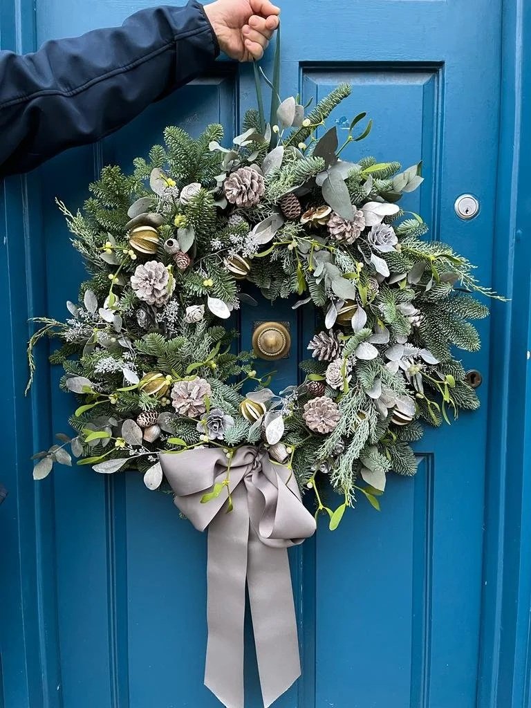A holiday wreath made of greenery, pinecones, and flowers, with a grey ribbon bow, hanging on a blue door, being held by a person's hand.