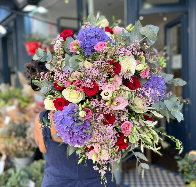 Person holding a large bouquet of mixed flowers including purple hydrangeas, red roses, white roses, pink lisianthus, and eucalyptus and olive leaves and foliage, in a floral, florist shop.