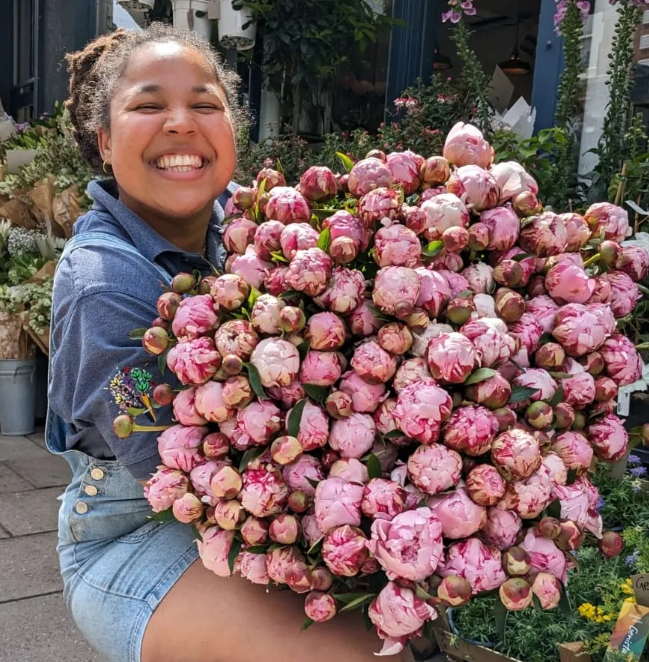 A young black woman with a big smile holding a large bouquet of pink peonies outside a florist shop