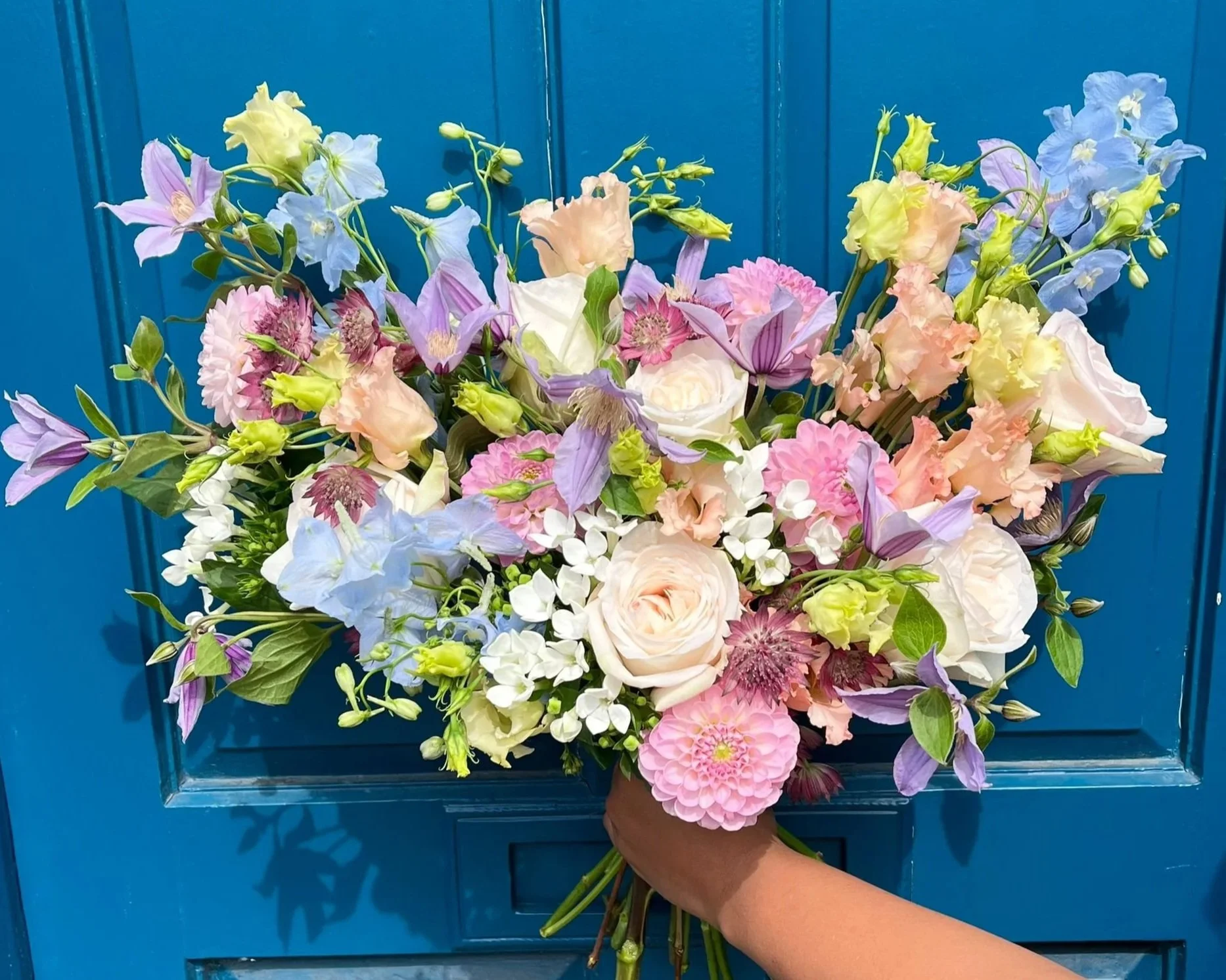 A hand holding a large bouquet of mixed pastel-colored flowers against a blue background.