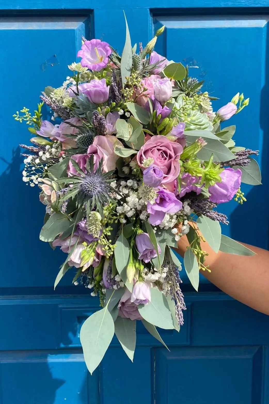 A vibrant bouquet of purple, pink, and white flowers with greenery, held in front of a blue wooden door.