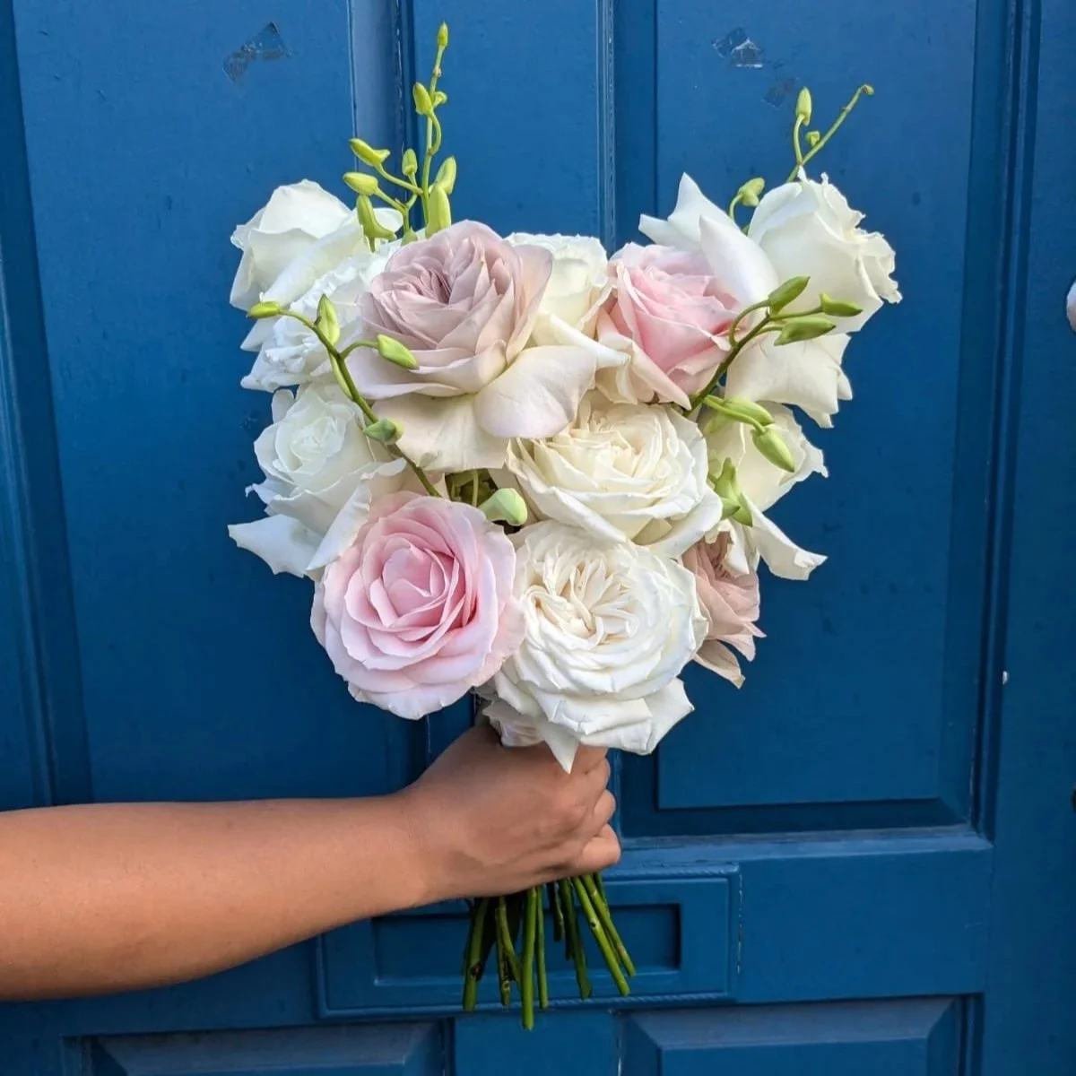 A bouquet of light pink and white roses with white and green buds, held by a person's hand in front of a blue door.