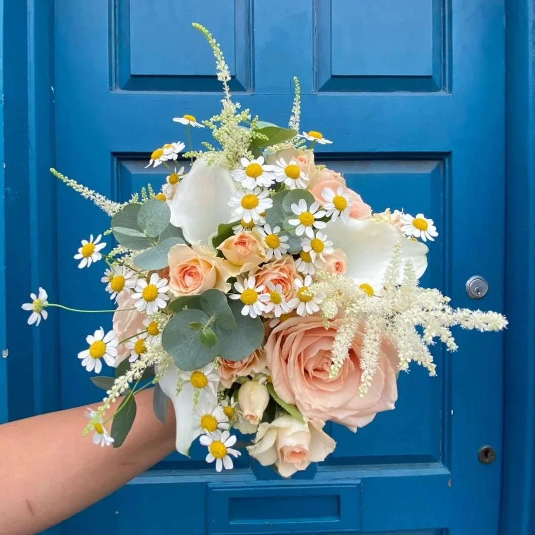 A floral bouquet featuring peach roses, white calla lilies, daisies with yellow centers, and eucalyptus leaves, held in front of a blue door.