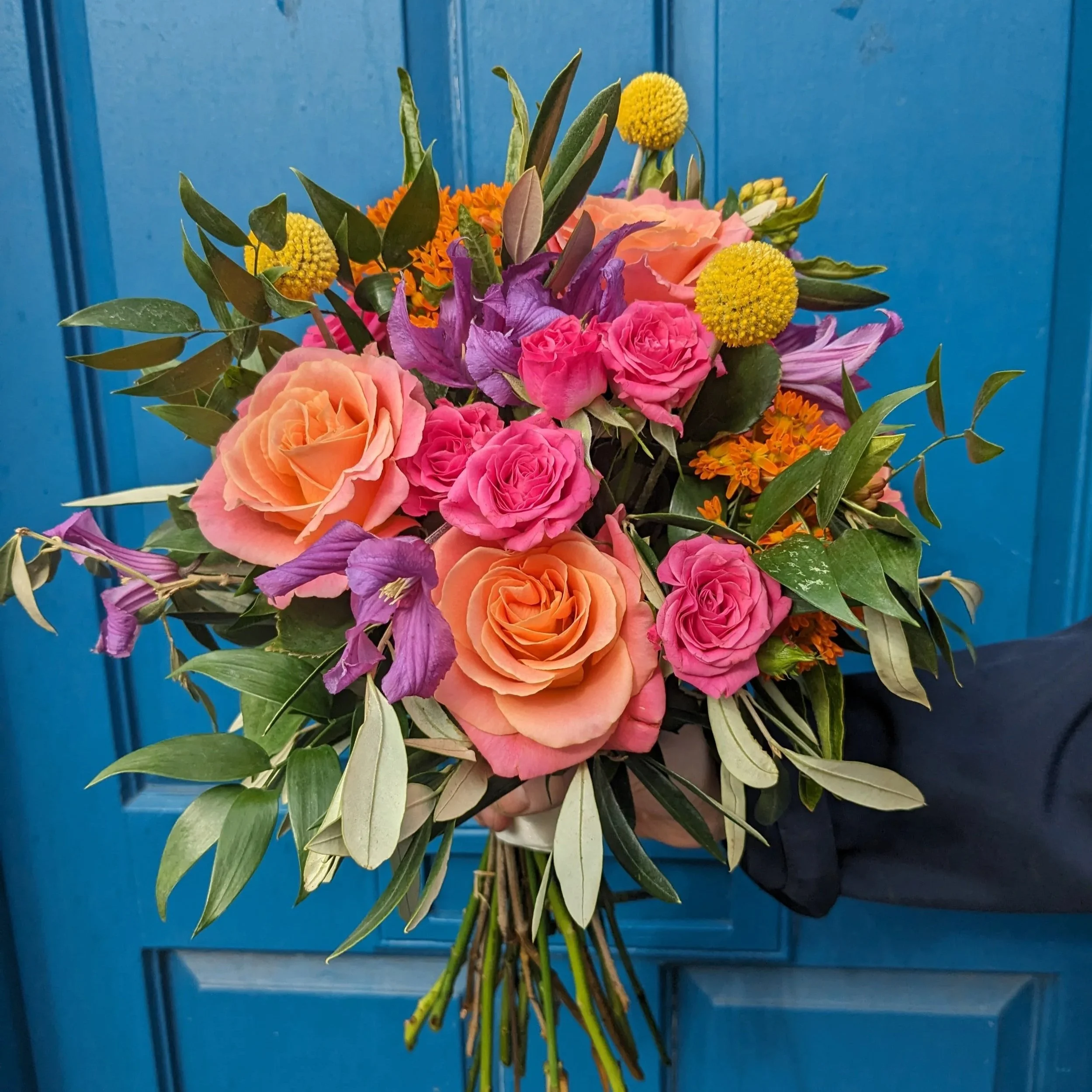 A bouquet of mixed flowers held by a person against a blue wooden background. The bouquet includes pink roses, purple clematis, yellow billy balls craspedia, orange blooms ascelpia, and green olive leaves.