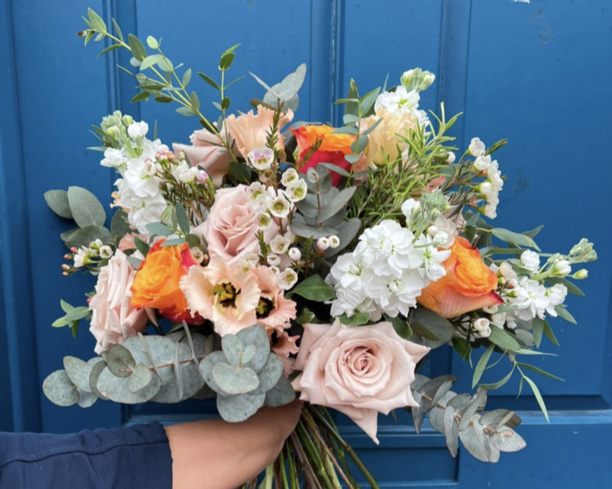 A person holding a bouquet of mixed flowers including blush roses, orange roses, white stock flowers, and eucalyptus against a blue background.