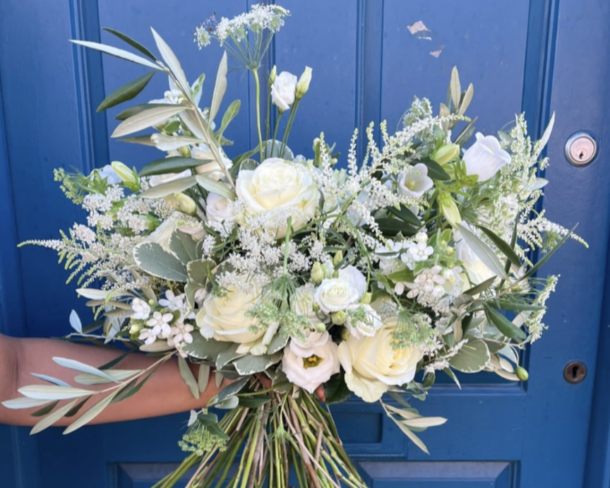 A bouquet of white roses, campanula, and various white flowers and greenery held against a blue door.