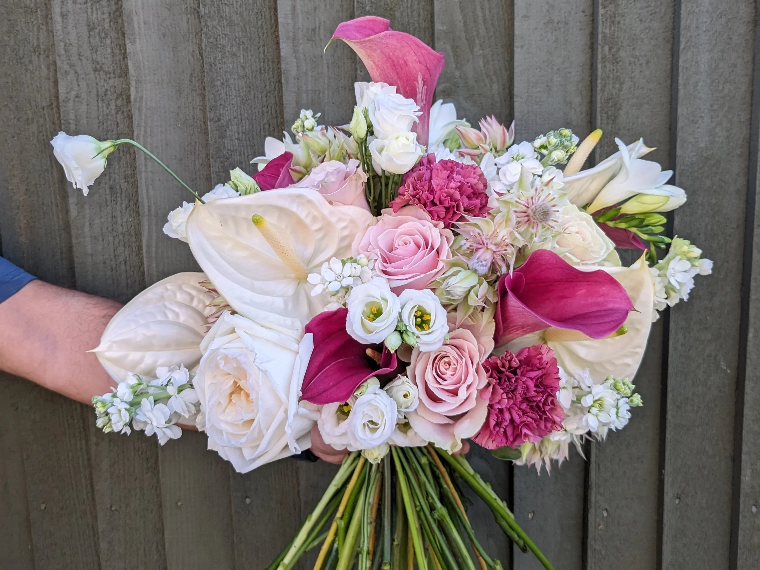 A colorful bouquet of various flowers including white calla lilies, pink roses, pink carnations, and other white and pink blossoms laid on a dark wooden surface.