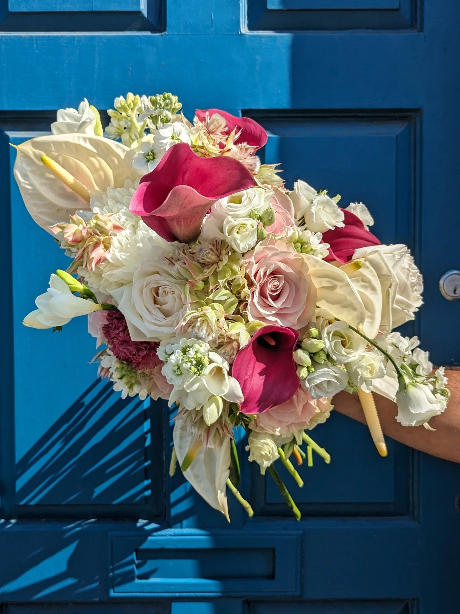 A colorful bouquet of flowers including white roses, pink calla lilies, and other various white and green flowers, held against a blue panel door with shadows from sunlight.