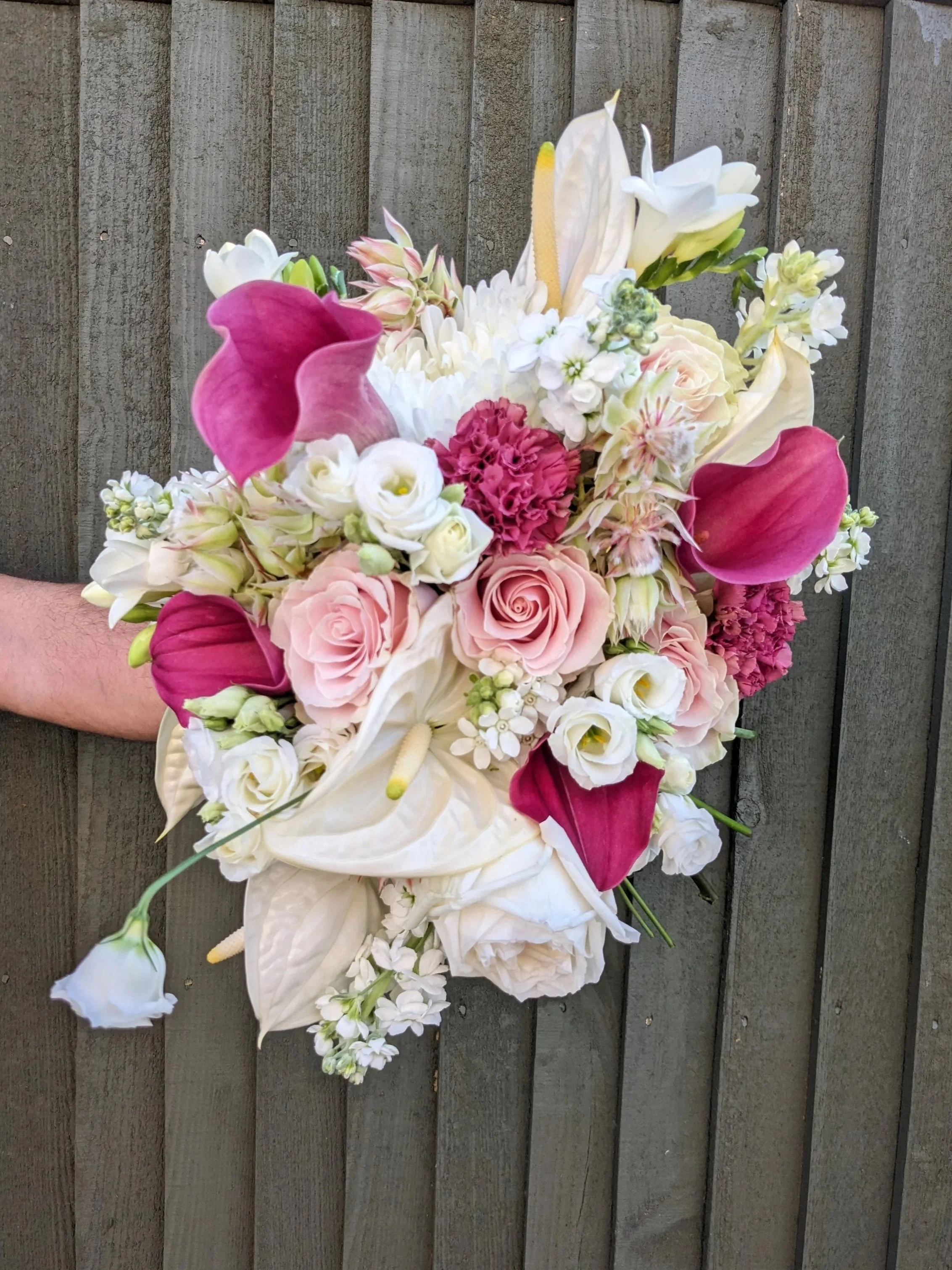 A bouquet of mixed flowers including pink calla lilies, white roses, and other white and cream flowers, arranged against a wooden background.