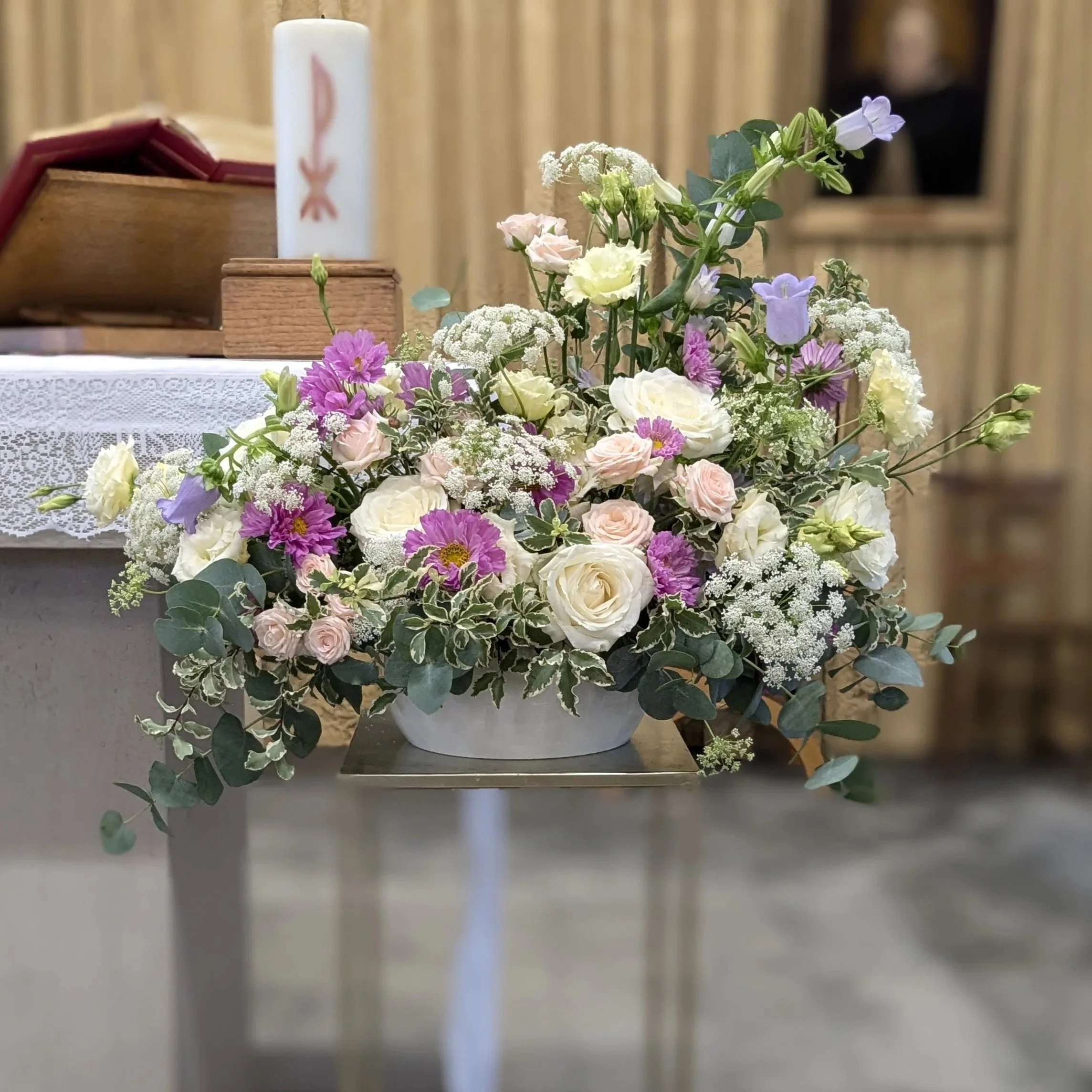 A bouquet of white, pink, purple, and cream flowers with green leaves and stems, arranged on a white lace tablecloth. In the background, a white candle with pink designs and a wooden box with a burgundy item inside are visible. The setting appears to